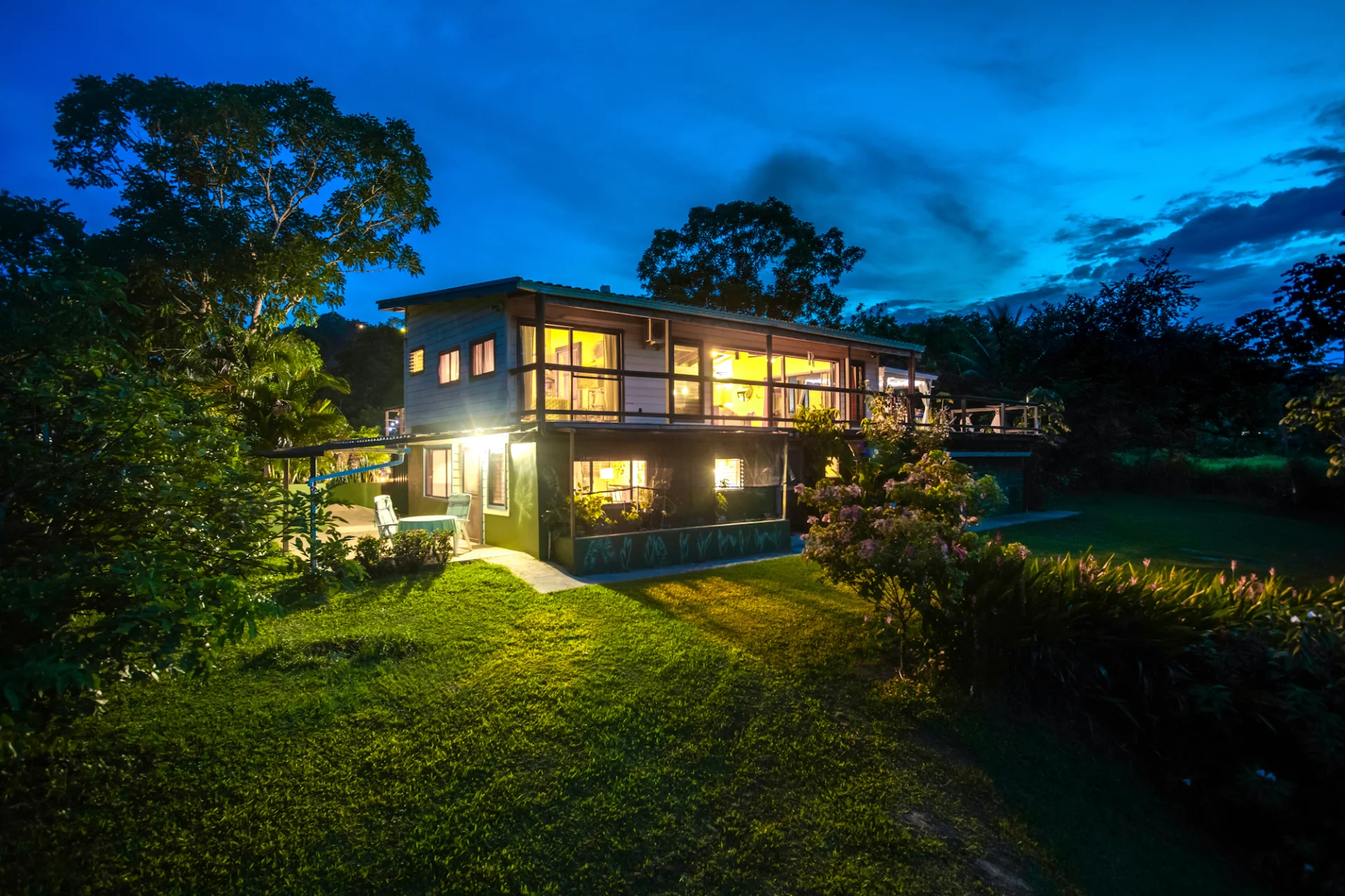 A warmly lit inland home in Belize at dusk, surrounded by lush tropical gardens